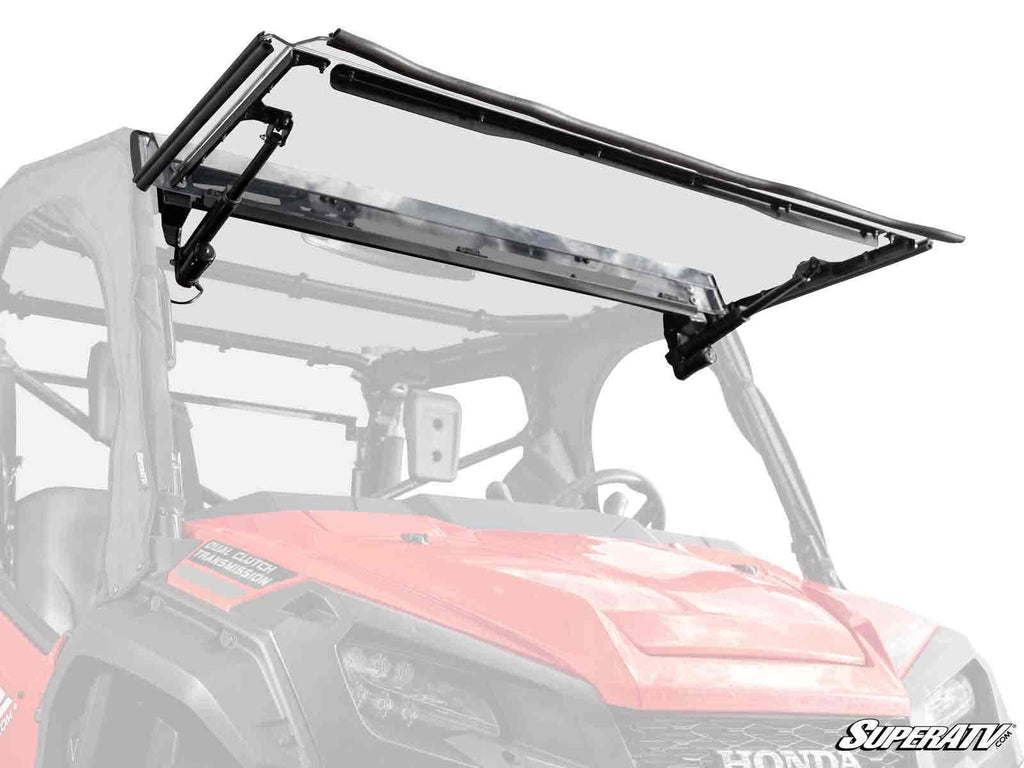 A red Honda side-by-side UTV features a Honda Pioneer 1000 MaxDrive Power Flip Windshield, shown raised with gas struts and black brackets. The SuperATV logo is in the lower right; frame and side mirrors are partly visible.