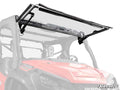 A red Honda side-by-side UTV features a Honda Pioneer 1000 MaxDrive Power Flip Windshield, shown raised with gas struts and black brackets. The SuperATV logo is in the lower right; frame and side mirrors are partly visible.
