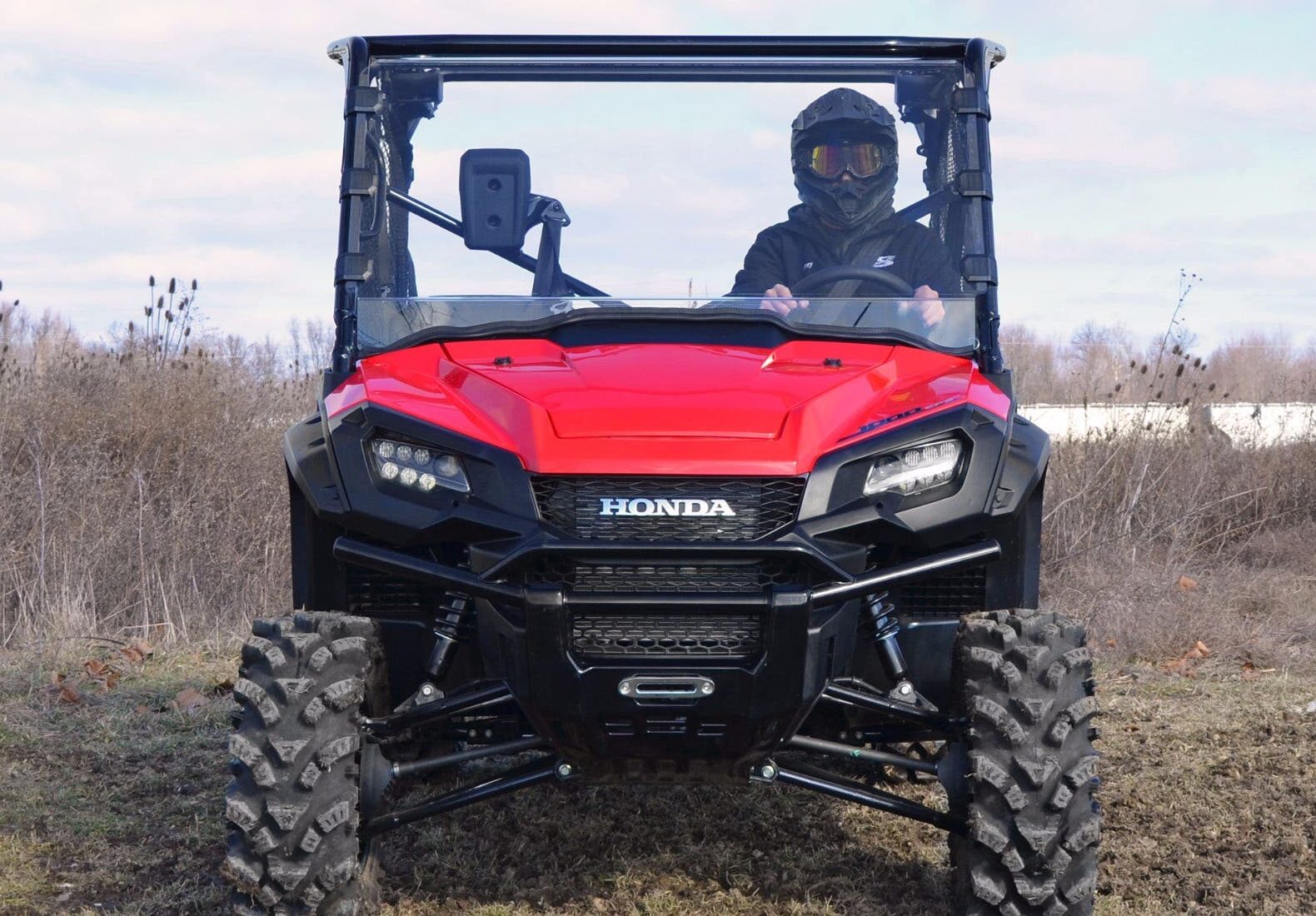 A person in a helmet and dark clothing sits facing the camera in a red Honda equipped with a Pioneer 1000 Scratch Resistant Full Windshield, parked on grass with rugged tires, dry vegetation, and a cloudy sky in the background.