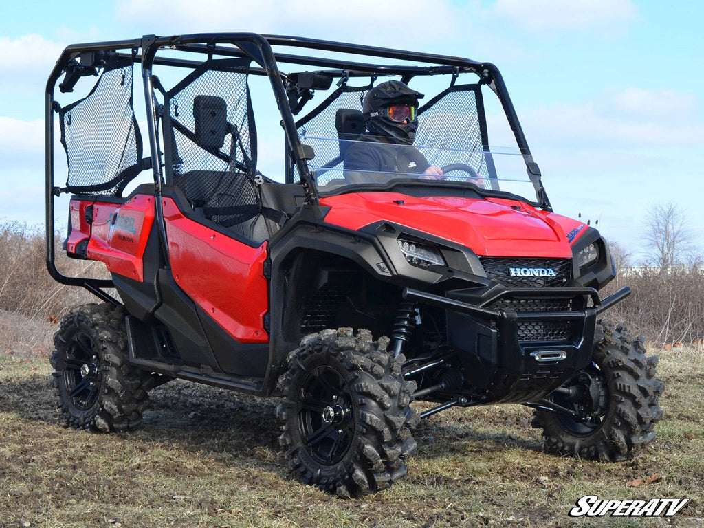 A person wearing a helmet sits inside a red Honda off-road utility vehicle with large tires and mesh doors, equipped with the Honda Pioneer 1000 Half Windshield, parked on rough grass under a blue sky.