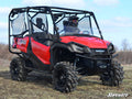 A person wearing a helmet sits inside a red Honda off-road utility vehicle with large tires and mesh doors, equipped with the Honda Pioneer 1000 Half Windshield, parked on rough grass under a blue sky.