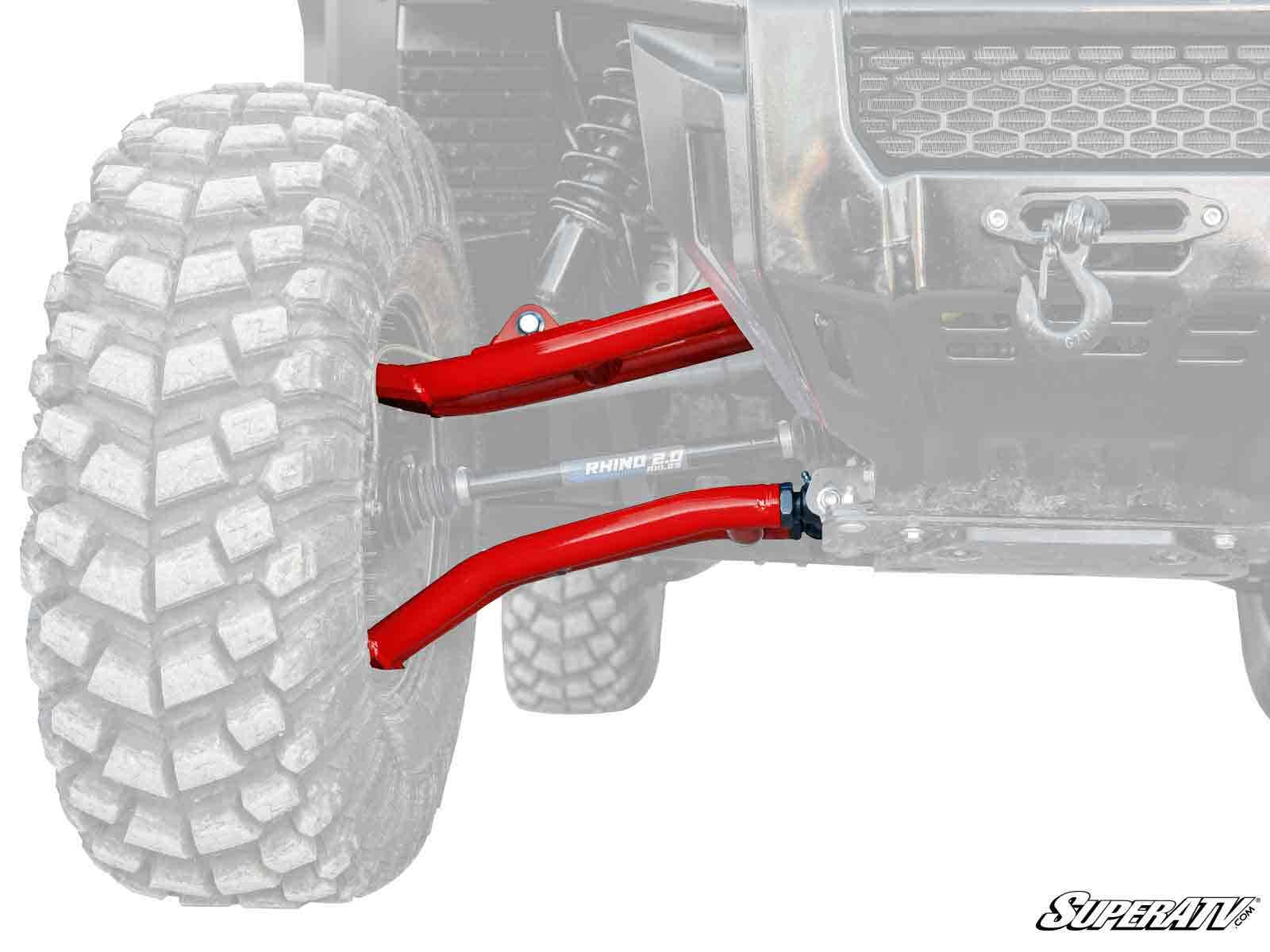 Close-up of the Honda Pioneer 1000 Atlas Pro 1.5 Forward Offset A-Arms, shown in red on an off-road vehicle. The rugged tire tread stands out, while the SuperATV logo appears in the corner and the rest of the vehicle is faded for emphasis.