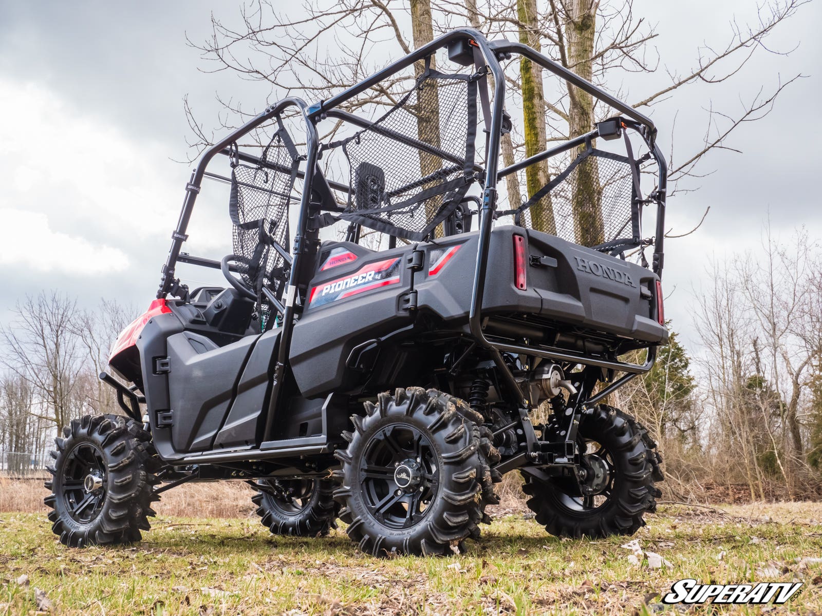 A Honda Pioneer 700 with a 2 Lift Kit, large off-road tires, and a protective cage is parked on grass. Trees and cloudy skies are in the background. The vehicle is viewed from the rear driver’s side with “SuperATV” at the bottom right.