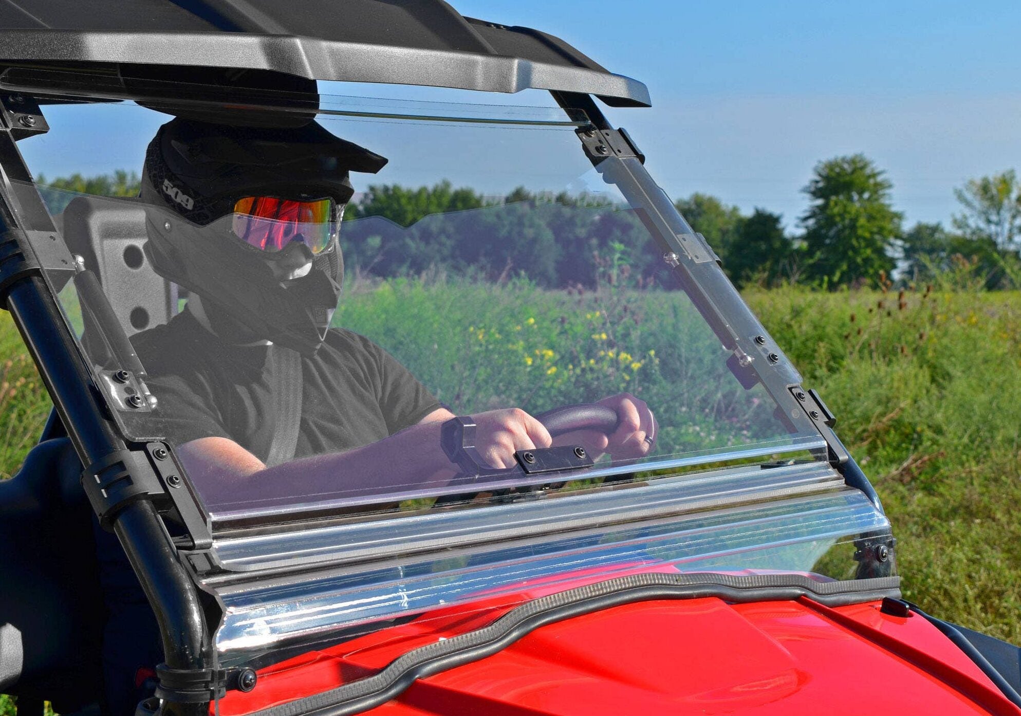 Wearing a black helmet and reflective goggles, a driver navigates a grassy landscape in a red vehicle equipped with the Honda Pioneer 500 Flip Windshield, surrounded by trees and wildflowers under a blue sky.