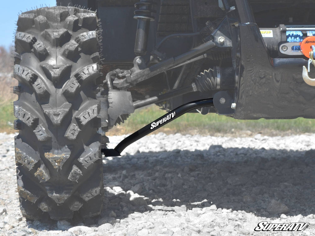 Close-up of an off-road vehicles front left suspension and wheel on rocky terrain. The Honda Pioneer 700 High Clearance A-Arms by SuperATV are clearly visible, featuring robust hardware and a thick treaded tire. SuperATV logo shown at lower right.