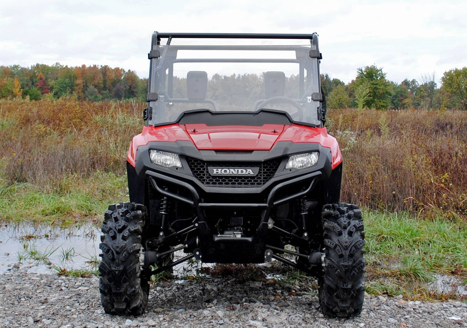 A Honda Pioneer 700 featuring a scratch-resistant full windshield is parked on rocky ground. It has large, rugged tires and a roll cage, with autumn trees and grassy fields visible under a cloudy sky in the background.