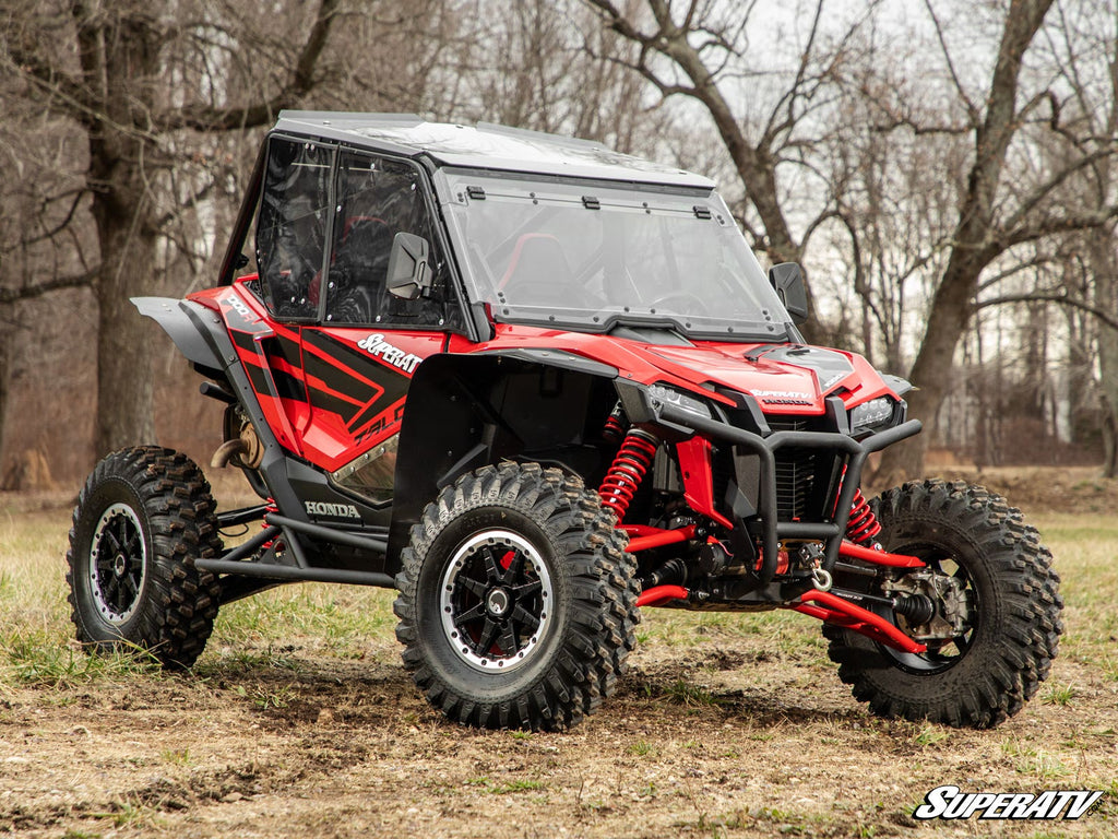 A red and black Honda Talon with large rugged tires and SuperATV branding is parked on grass. It features a MaxDrive Power Flip Windshield with clear doors, against a backdrop of leafless trees and an overcast sky.