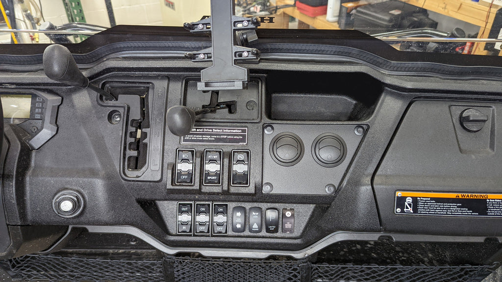 Dashboard of a Honda Pioneer 1000 (2022-2024) with an Ice Crusher Cab Heater, showing gear levers, control switches, toggle switches, circular dials, a warning label, and a device mount above the steering controls; workshop shelves/tools in background.