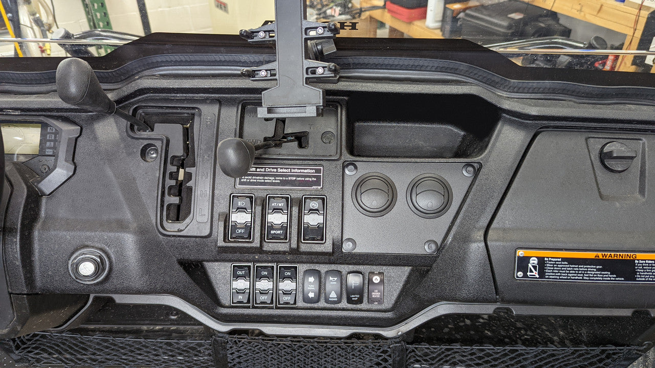 Dashboard of a Honda Pioneer 1000 (2022-2024) with an Ice Crusher Cab Heater, showing gear levers, control switches, toggle switches, circular dials, a warning label, and a device mount above the steering controls; workshop shelves/tools in background.