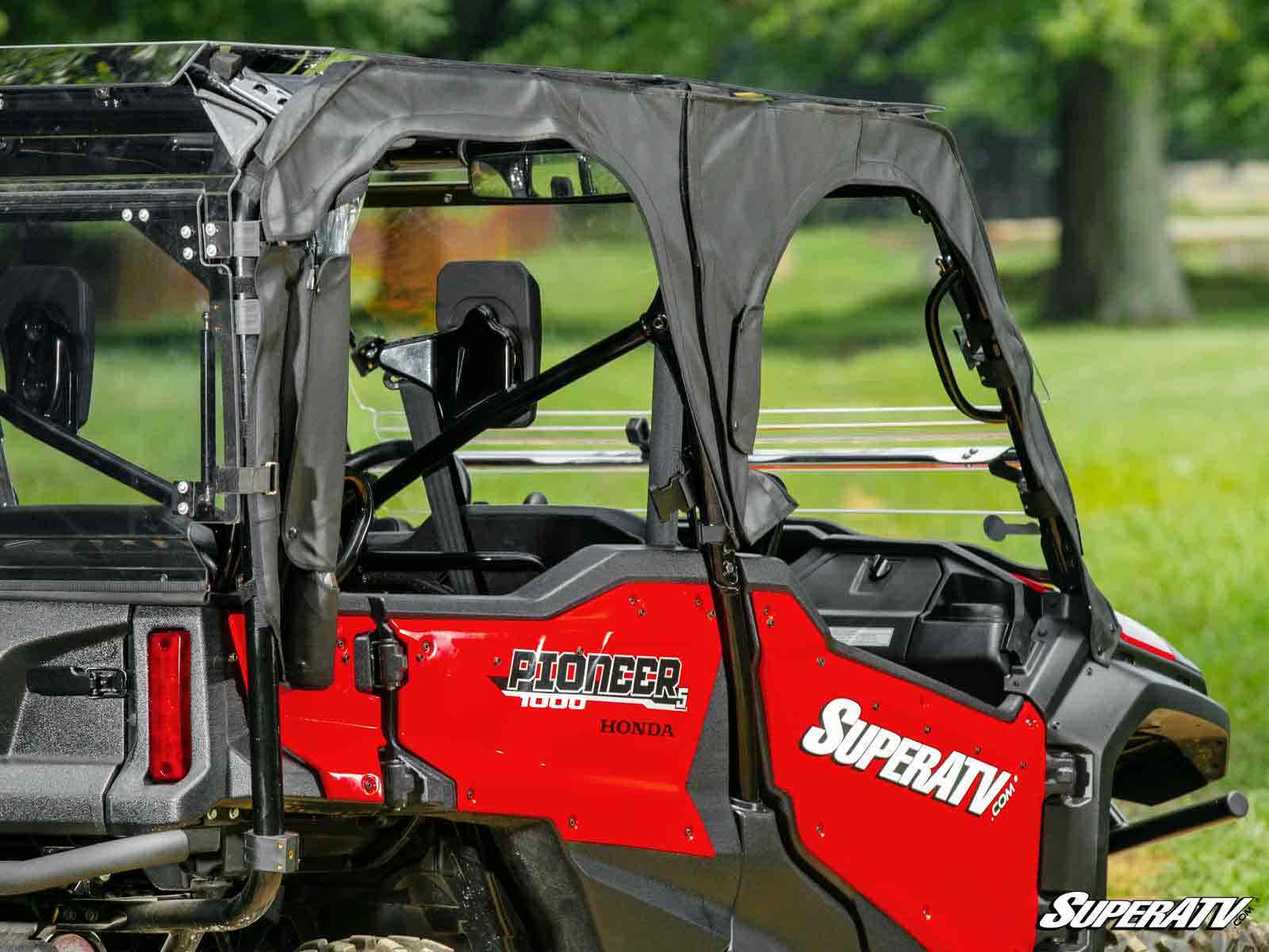 A close-up of a red Honda Pioneer 1000-5 with Primal Soft Cab Enclosure Upper Doors, featuring clear windows and SuperATV branding, parked on grass with trees blurred in the background.