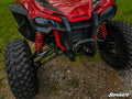 A close-up of the Honda Talon 1000R High Clearance A-Arms, front suspension, and rugged tires of a red off-road vehicle parked on gravel with nearby grass. Large coil springs, control arms, and part of the grill are visible. SuperATV logo at bottom right.