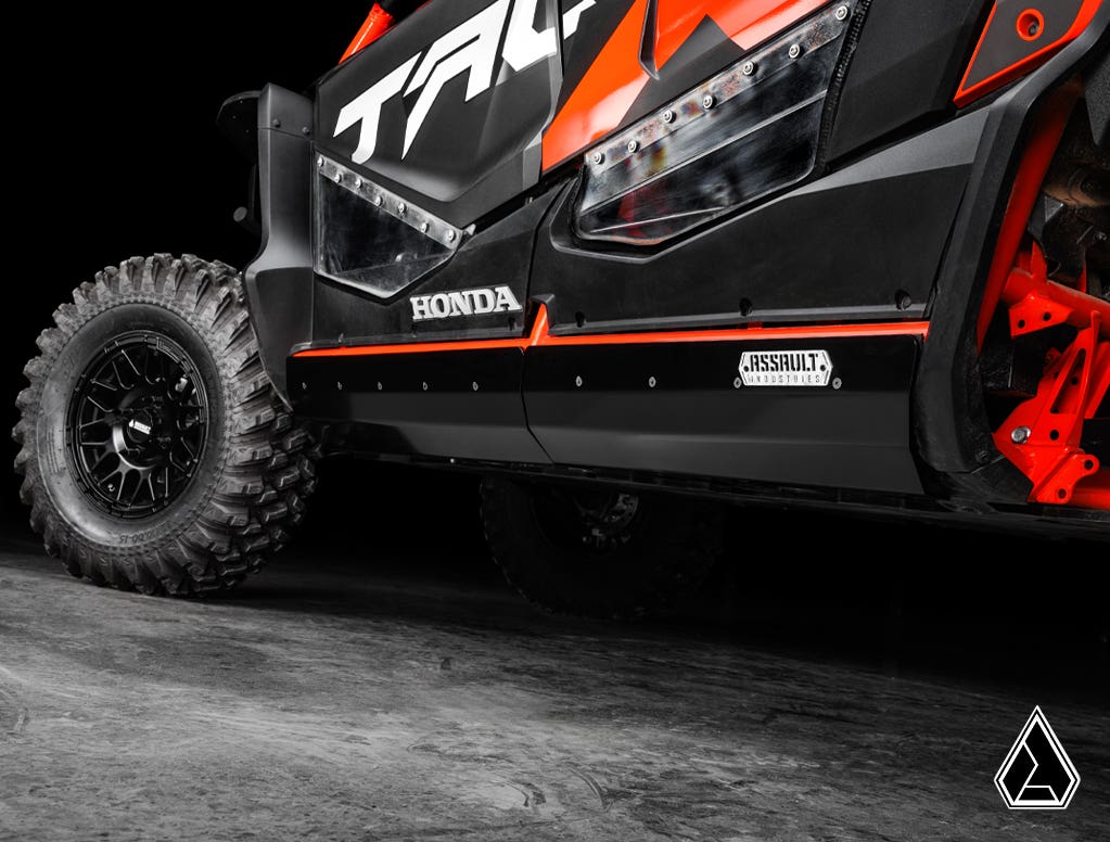 Close-up of Assault Industries Honda Talon 1000X-4 Rock Sliders installed on a black and red off-road vehicle with rugged tires and angular body panels, visible logos, on a concrete floor against a dark background.