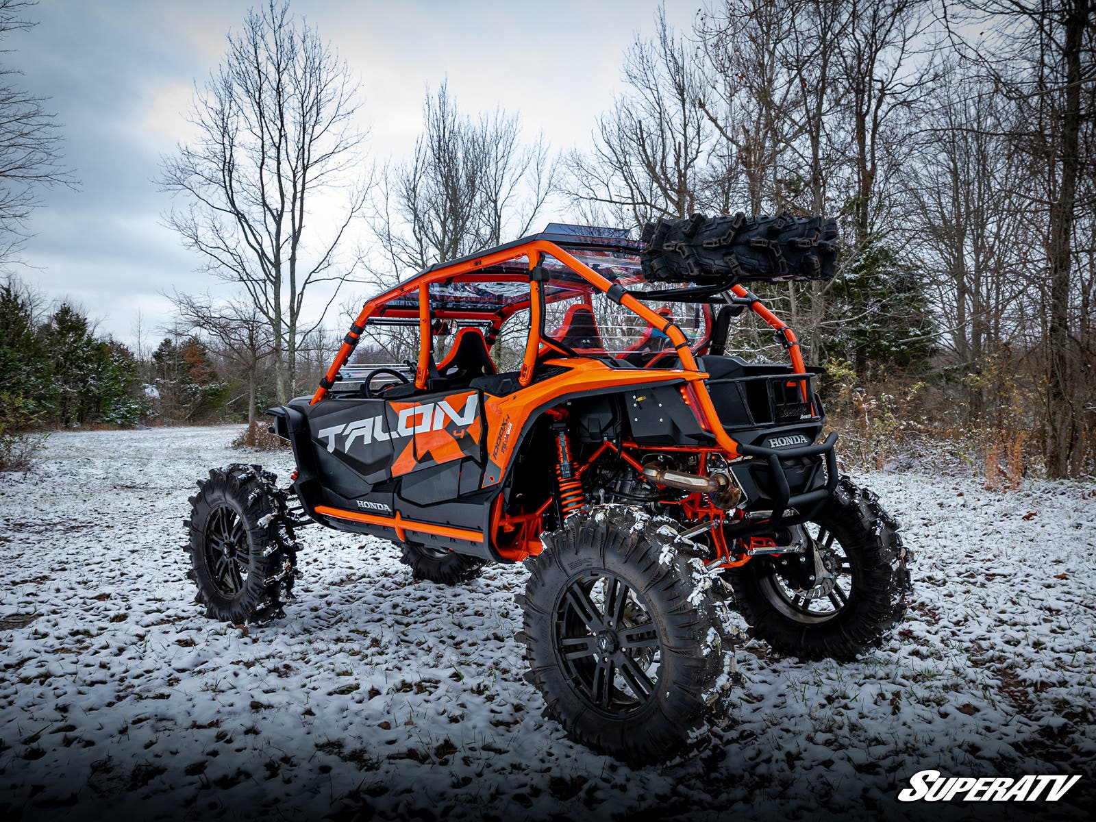 A Honda Talon 1000 off-road vehicle with a spare tire carrier is parked on snowy ground among bare trees. It features large tires, a roll cage, and Talon branding. The sky is cloudy and the ground has a light snow cover.