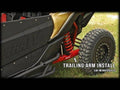 Close-up of a Can-Am Maverick X3 with 72 Rear Trailing Arms, highlighting the red trailing arm, coil spring suspension, rugged tire, and black side panel on grass. Text: TRAILING ARM INSTALL – CAN-AM MAVERICK X3 72.
