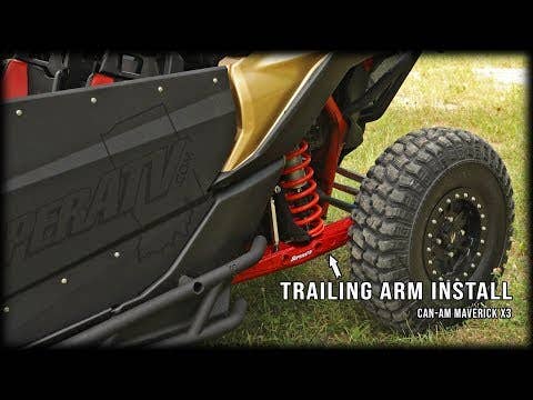 Close-up of a Can-Am Maverick X3 with 72 Rear Trailing Arms, highlighting the red trailing arm, coil spring suspension, rugged tire, and black side panel on grass. Text: TRAILING ARM INSTALL – CAN-AM MAVERICK X3 72.