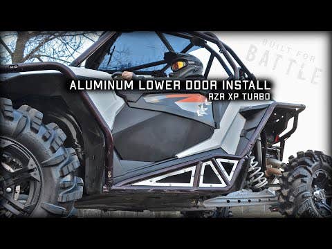 A person wearing goggles sits in a rugged black and gray RZR XP Turbo with large knobby tires. The image highlights the installation of Polaris RZR XP Turbo Lower Doors. Bold text reads: ALUMINUM LOWER DOOR INSTALL RZR XP TURBO.