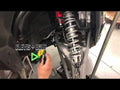 A hand points at the Assault Industries Polaris RZR XP Turbo S Turret-Style Heavy-Duty Tie Rods inside a vehicle’s wheel well, highlighting the CLEVIS + BOLT area with a green triangle; tie rods and suspension components are visible.