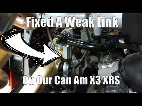 Close-up of Assault Industries Barrel Rear Sway Bar End Links (Fits: Can-Am Maverick X3) with a white arrow pointing to the upgraded link. Overlay: Fixed a Weak Link on Our Can-Am X3 XRS. Engine and suspension parts are visible nearby.