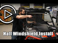 A man installs a Can-Am Defender Half Windshield on a black off-road vehicle in a garage. Text reads Half Windshield Install with a large play button on the left. Shelves and vehicle parts can be seen in the background.