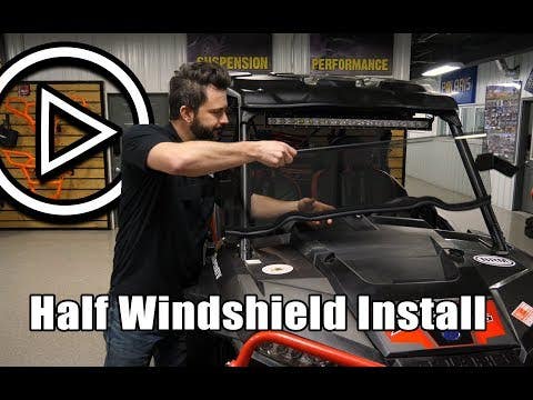 A man installs a Gravely Atlas JSV Half Windshield on an off-road vehicle in a garage, with shelves of auto parts behind him. A large play button is on the left and bold text at the bottom reads, “Half Windshield Install.”.