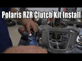 A person installs a Polaris RZR 900 Clutch Kit on a workbench, using a power tool on the clutch assembly. Large white text at the top reads, “Polaris RZR 900 Clutch Kit Install,” with various tools and parts in the background.