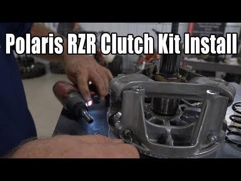 Close-up of hands installing a Polaris Ranger XP 1000 Clutch Kit. One hand uses a tool while the other steadies the clutch assembly. Workshop setting with Polaris Ranger XP 1000 Clutch Kit Install in bold at the top.