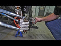 A person works on a Polaris RZR XP Turbo elevated on jack stands, inspecting its suspension with billet aluminum radius arms visible. Control arms, coilover shocks, and a brake caliper are exposed. Floor text reads “BUILT FOR BATTLE.” Tools and a door app