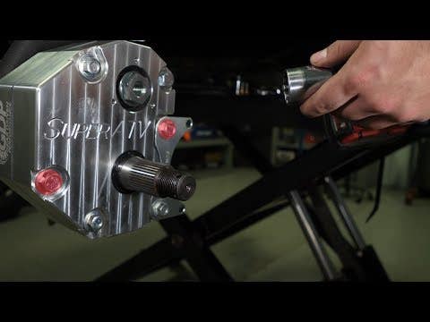 A close-up shows a person’s hand using a power tool on a Polaris RZR XP 1000 4 Portal Gear Lift. The cylindrical part extends from the gear lift, which is mounted on a black frame in a workshop setting.
