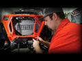 A man in a red shirt and cap installs or tests wiring on the front of an orange off-road vehicle inside a garage, where the grill is removed to expose the radiator and a Polaris RZR XP Turbo S Winch Mount.