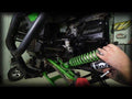 A person installs a Kawasaki Teryx 2 Lift Kit onto an off-road vehicles suspension with a black SuperATV panel visible in a workshop setting. Various mechanical parts are exposed during the installation process.