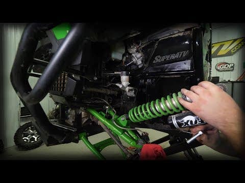 A person installs a Kawasaki Teryx 2 Lift Kit onto an off-road vehicles suspension with a black SuperATV panel visible in a workshop setting. Various mechanical parts are exposed during the installation process.