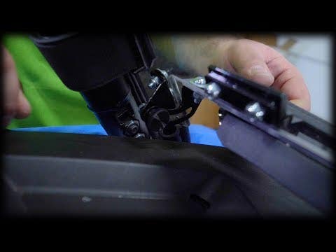 Close-up of hands installing the Polaris RZR XP 1000 Vented Full Windshield, securing it onto a black surface with visible screws and hardware. A blue cloth lies underneath, while a blurred background emphasizes the windshield installation process.