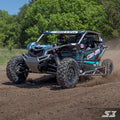 A black and teal Can-Am Maverick X3 with S3 Power Sports 2017+ Tree Kickers speeds across muddy terrain, kicking up dust. The helmeted driver navigates through dense green trees on a sunny day, showcasing rugged tires in action.