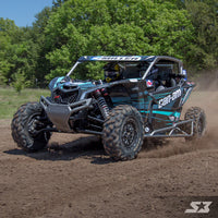 A black and teal Can-Am Maverick X3 with S3 Power Sports 2017+ Tree Kickers speeds across muddy terrain, kicking up dust. The helmeted driver navigates through dense green trees on a sunny day, showcasing rugged tires in action.