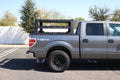 A 2009-2014 Ford F-150 with Treck - Overland Package (adjustable bed rack system, texture black) is parked in a lot. The sturdy rack is installed in the bed; trees and a white wall are visible under a clear sky.