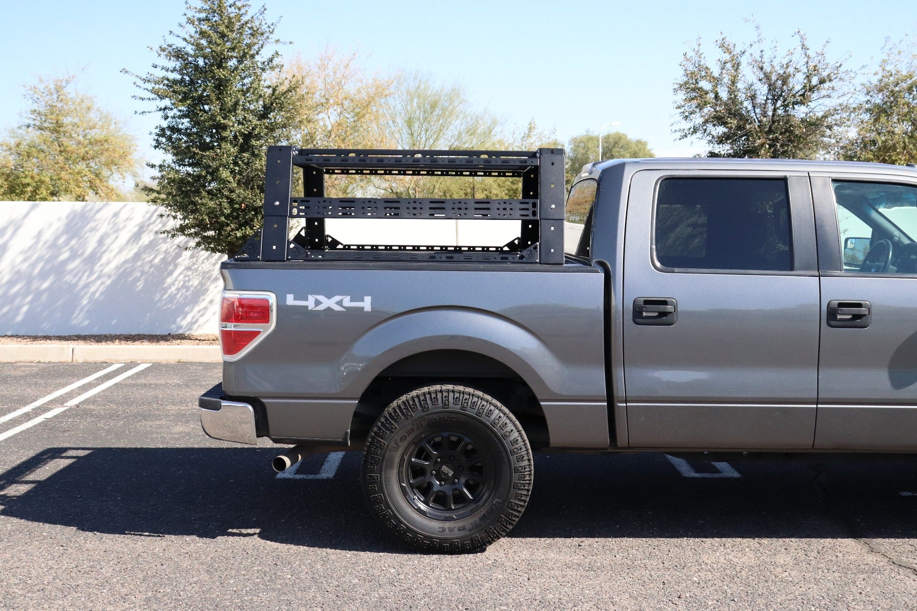A 2009-2014 Ford F-150 with Treck - Overland Package (adjustable bed rack system, texture black) is parked in a lot. The sturdy rack is installed in the bed; trees and a white wall are visible under a clear sky.