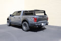A gray Ford F-150 with a Treck Overland Package bed rack system (texture black, adjustable, for 5½-6ft beds, with bracket kit and rails) is parked on black pavement near a beige brick wall under bright sunlight.