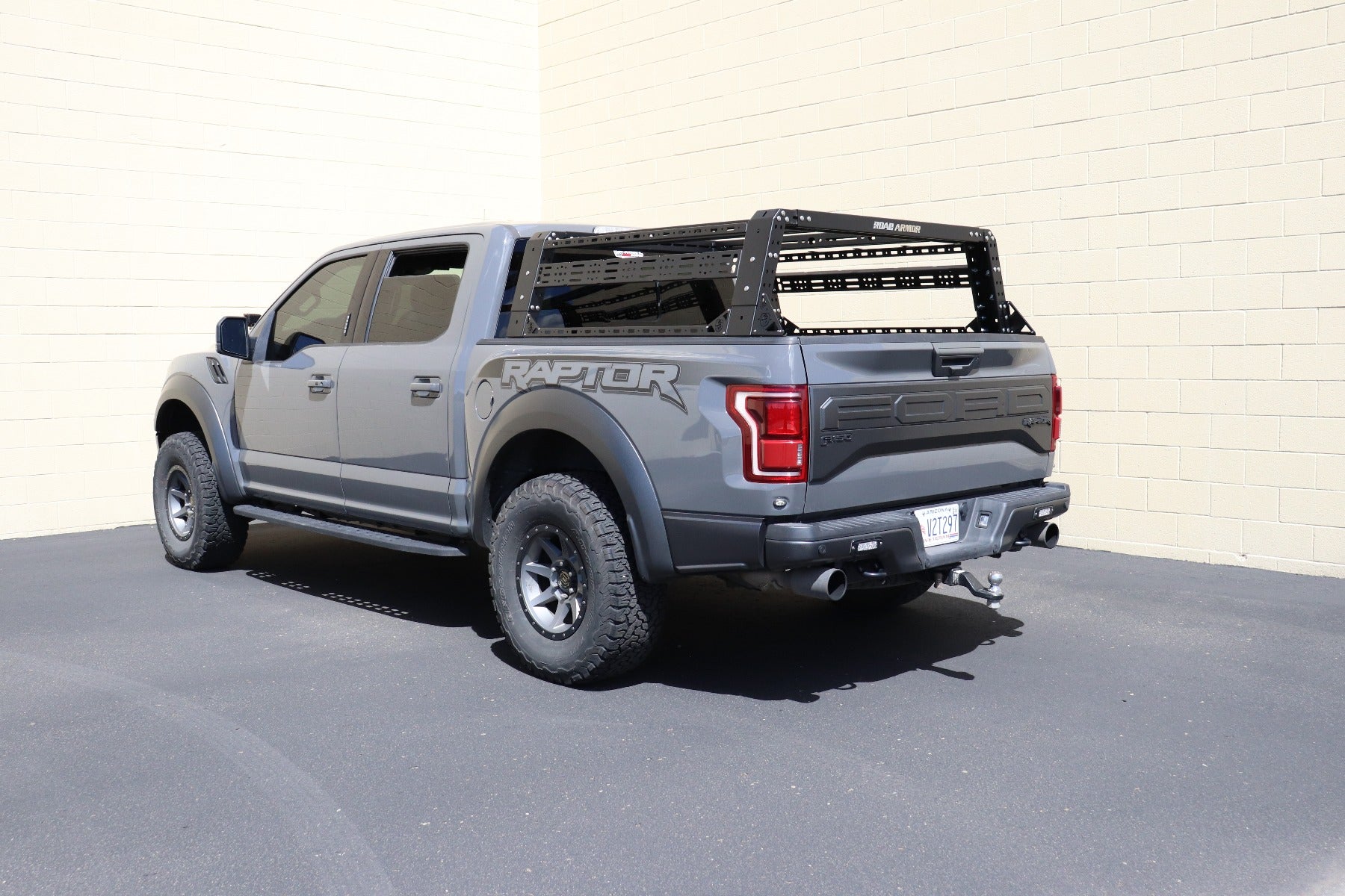 A gray Ford F-150 with a Treck Overland Package bed rack system (texture black, adjustable, for 5½-6ft beds, with bracket kit and rails) is parked on black pavement near a beige brick wall under bright sunlight.