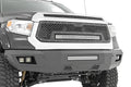 Close-up of a rugged Toyota Tundra (2014-2021) with a Rough Country Front Bumper, showcasing a black and silver finish, heavy-duty design, exposed suspension parts, large grille with metal rivets, and integrated LED light bars.