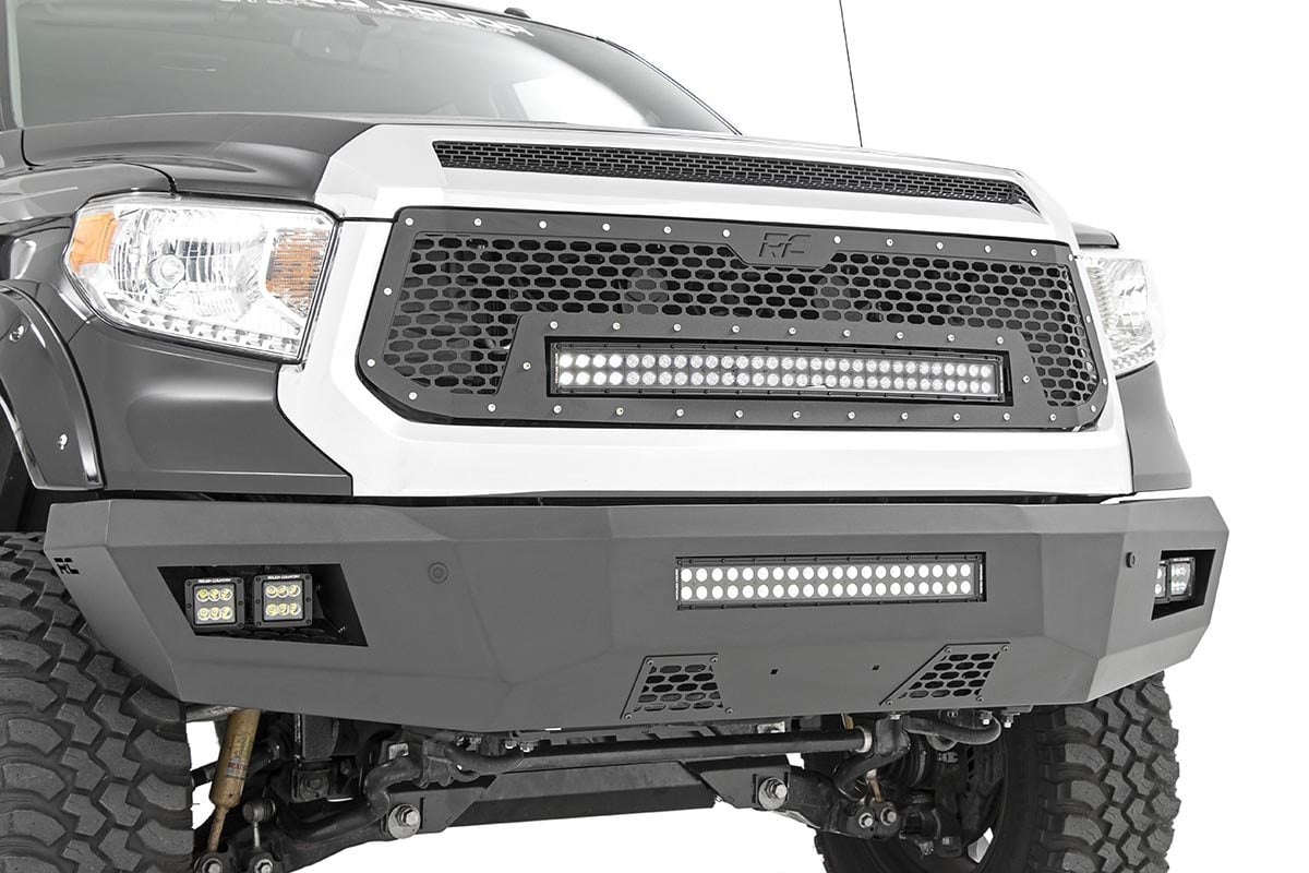 Close-up of a rugged Toyota Tundra (2014-2021) with a Rough Country Front Bumper, showcasing a black and silver finish, heavy-duty design, exposed suspension parts, large grille with metal rivets, and integrated LED light bars.