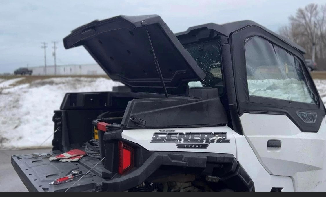 A JEMCO UTV Rear Cargo Box is shown installed on a Polaris General with its lid open. The vehicle is parked outdoors on snowy ground, with utility poles and an overcast sky in the background.