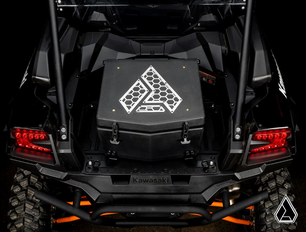 A rear view of a black Kawasaki Teryx KRX 1000 shows the Assault Industries Cooler/Cargo Box, featuring a white geometric logo and secured on the cargo bed. Red tail lights, rugged tires, and protective bars highlight the vehicle’s robust design.