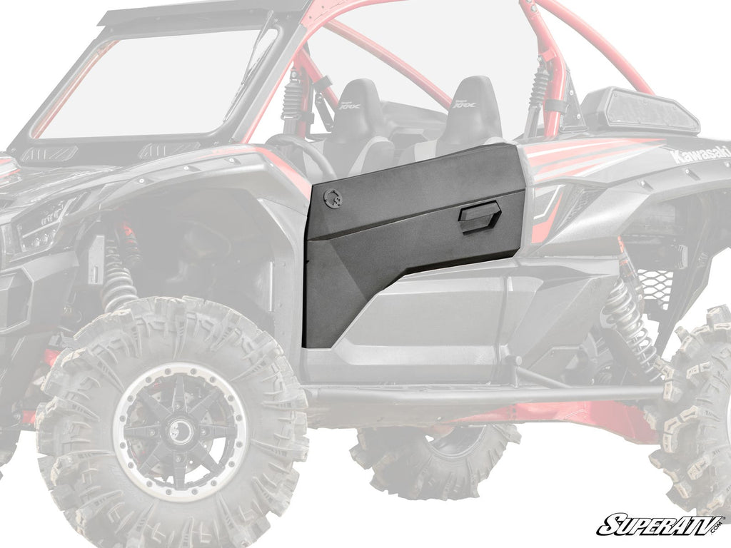 The Kawasaki Teryx KRX Aluminum Door Skin, shown in matte black with an angular design, is installed on the passenger side of a Kawasaki UTV. The door features a molded handle and distinct panel lines, standing out against the faded background vehicle.