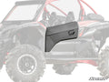 The Kawasaki Teryx KRX Aluminum Door Skin, shown in matte black with an angular design, is installed on the passenger side of a Kawasaki UTV. The door features a molded handle and distinct panel lines, standing out against the faded background vehicle.