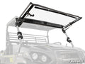 A Kawasaki Mule Pro Scratch Resistant Flip Windshield is installed on a rugged utility vehicle outdoors. The transparent windshield lifts on side support arms and mounts with brackets. The SuperATV logo appears in the lower right corner.