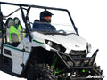 A person in a helmet and dark clothing sits in a white off-road side-by-side with green accents and rugged tires. The Kawasaki Teryx 800 Half Windshield by SuperATV is installed. The logo is visible against a fully white background.