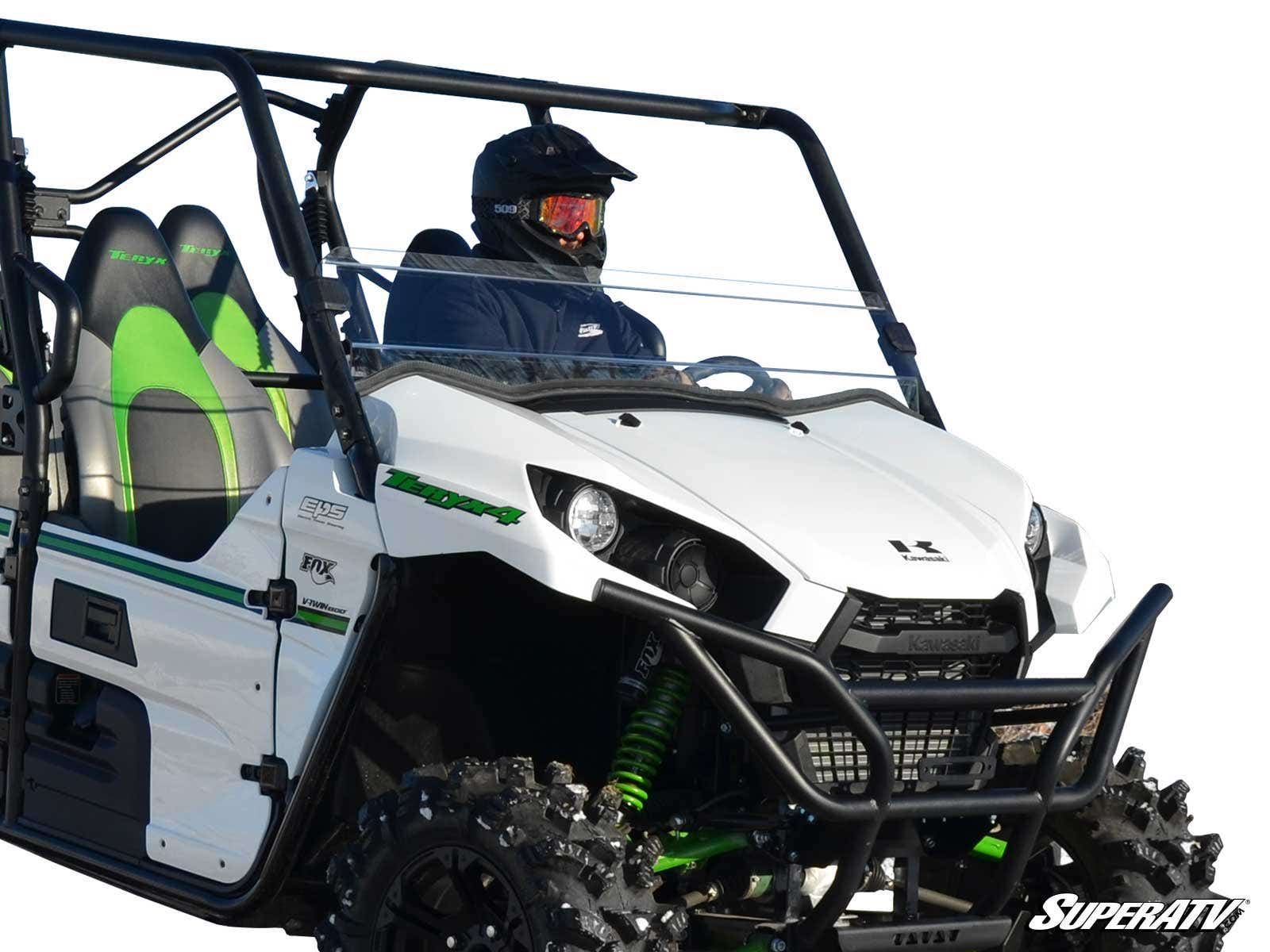 A person in a helmet and dark clothing sits in a white off-road side-by-side with green accents and rugged tires. The Kawasaki Teryx 800 Half Windshield by SuperATV is installed. The logo is visible against a fully white background.