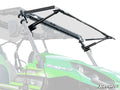 A close-up of a Kawasaki Teryx MaxDrive Power Flip Windshield shows the green off-road vehicle’s transparent windshield partially lifted on black hinges and supports, with the SuperATV logo visible in the bottom right corner.