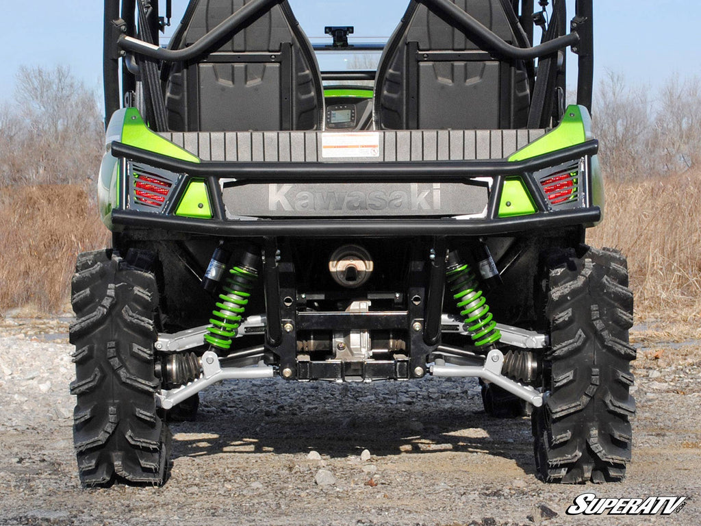 Rear view of a green Kawasaki Teryx 4 with its rear bumper, large rugged tires, bright green suspension springs, exposed undercarriage parts, and visible seats and harnesses; dried grass and a clear sky are in the background.