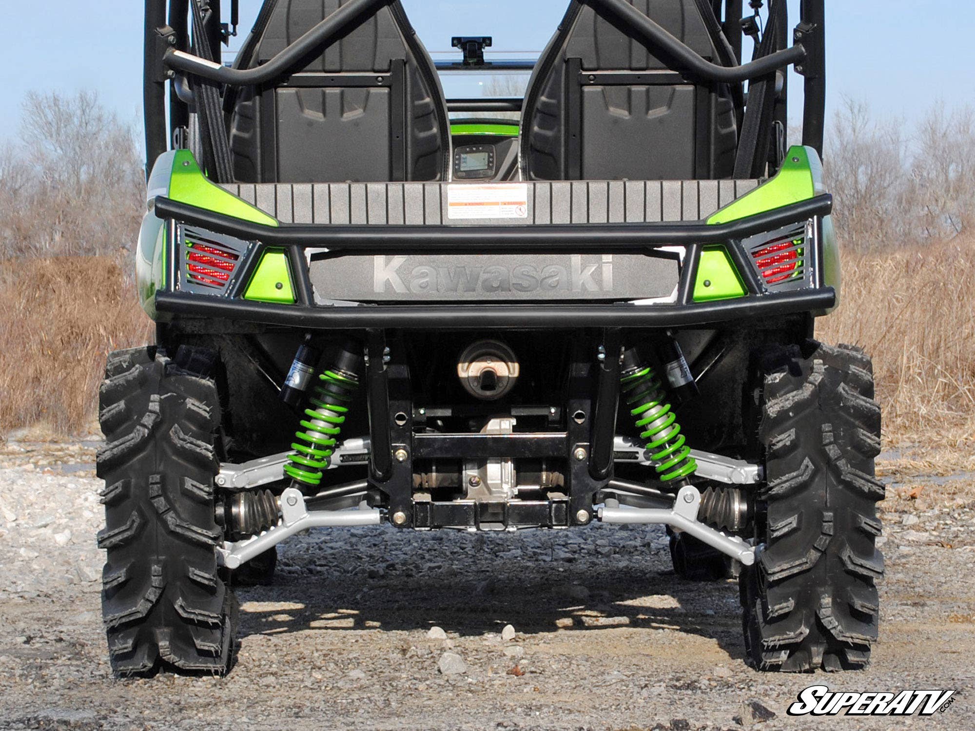 Rear view of a green Kawasaki Teryx 4 with its rear bumper, large rugged tires, bright green suspension springs, exposed undercarriage parts, and visible seats and harnesses; dried grass and a clear sky are in the background.